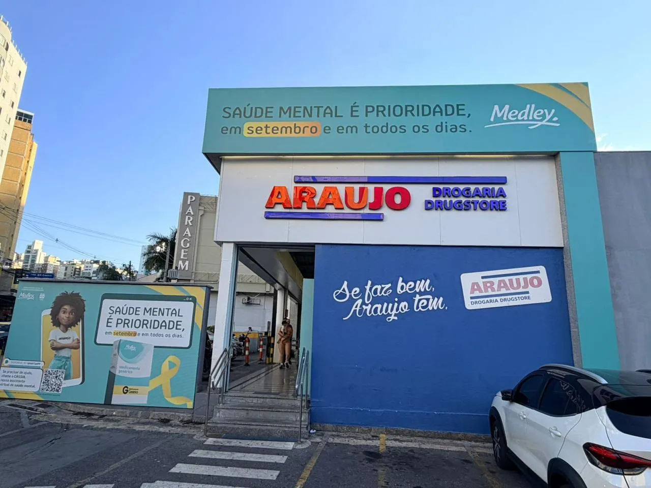 Fachada de drogaria com letras caixa, testeira verde-água com campanha impressa e painel lateral com ilustração