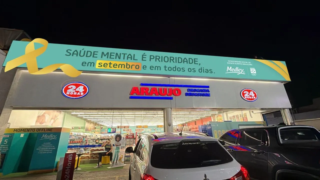 Fachada de drogaria à noite, com letras caixa iluminadas em vermelho e azul e testeira verde-água com campanha impressa