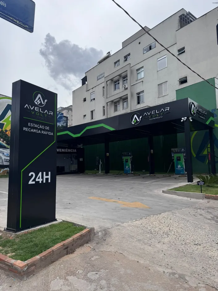 Eletroposto de dia, com totem preto "Estação de recarga rápida 24H" e cobertura com a marca Avelar Volt e faixa verde