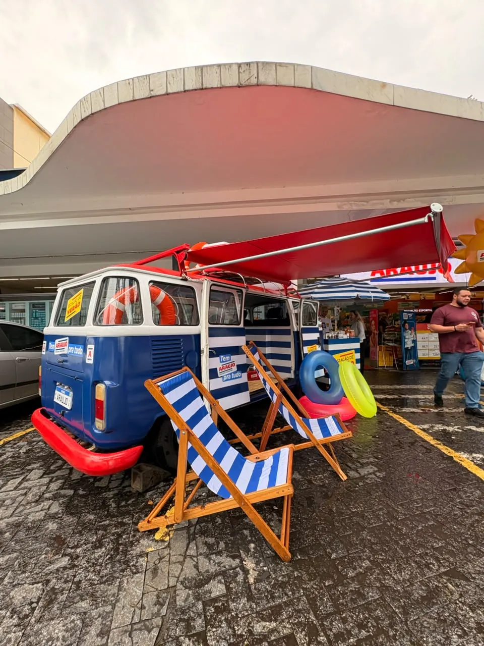 Kombi azul e branca com toldo vermelho, cadeiras de praia listradas e boias, montada em estacionamento