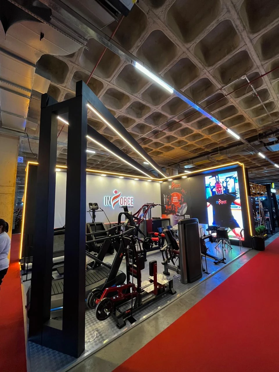 Estande em feira, com equipamentos de academia expostos sob iluminação laranja e telão de vídeo