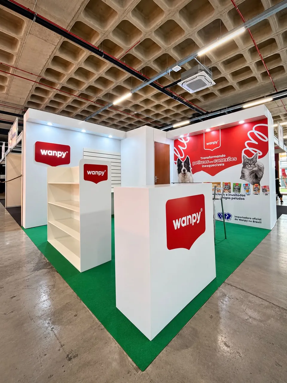 Estande branco e vermelho, com expositores, balcão e painel com cão e gato, sobre piso verde, em pavilhão de feira