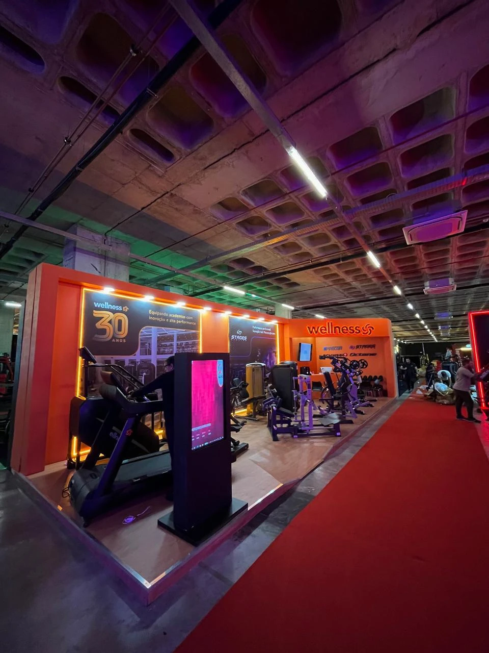 Estande laranja e branco, com esteira, aparelhos de musculação e totem digital, em pavilhão de feira
